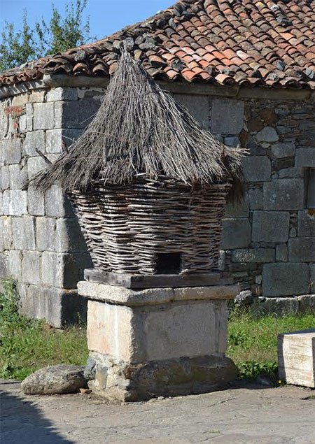 Un cabazo, rústico hórreo, en el centro de Leboreiro, La Coruña. Detrás se ve la sólida construcción de piedra del viejo hospital medieval. Imagen de José Holguera para Guiarte.com.